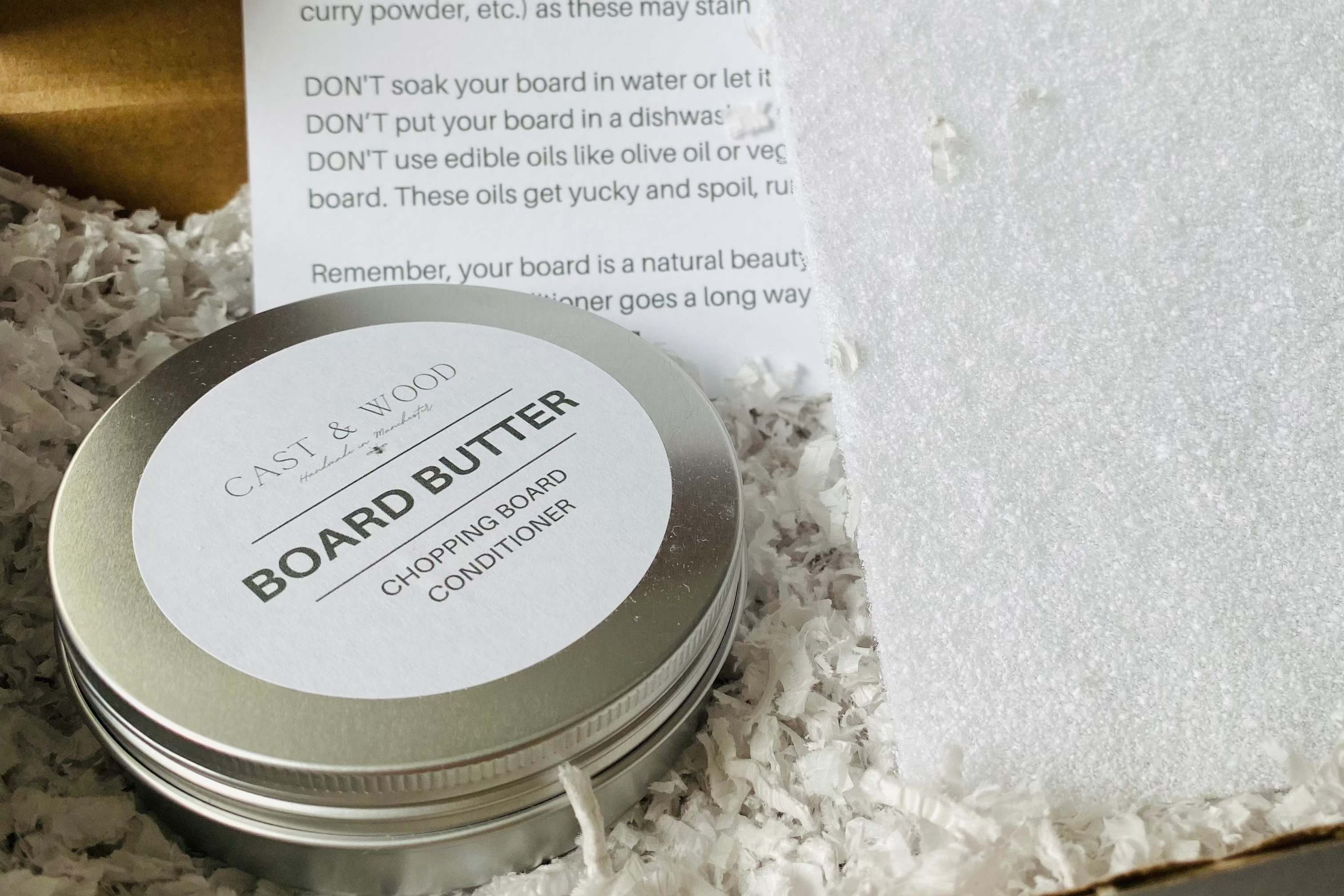 Round tin labelled 'BOARD BUTTER' for conditioning chopping boards, placed in a box with shredded paper and care instructions.